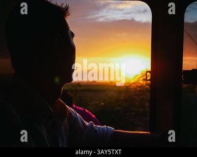 Ein Mann schaut nachdenklich aus einem Fenster und bewundert die atemberaubende Schönheit des Sonnenuntergangs, während er den Himmel mit leuchtenden Farben übermalt Stockfoto