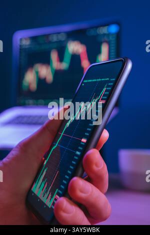 Nahaufnahme, Trader Hand mit Smartphone mit Finanzhandels-App, die volatile Trends bei Kryptowährungen und Börsen zeigt. Man kauft nachts Krypto auf seinem Smartphone in einem neonbeleuchteten Büro Stockfoto