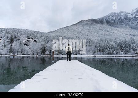 Ein Mädchen mit Rucksack steht auf einem verschneiten Dock mit Blick auf einen ruhigen See, umgeben von schneebedeckten Bäumen und einem majestätischen Berg. Stockfoto