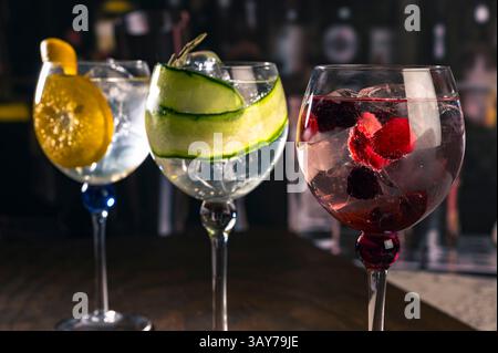 Drei stilvolle Cocktails, serviert in großen Gläsern mit Orange, Gurke und roten Beeren, stehen auf einer Holzoberfläche in einer stimmungsvollen Bar-Atmosphäre, ideal für Stockfoto