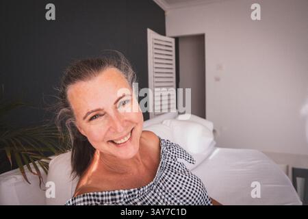 Ältere Frau, die auf einer weißen Couch in einem modernen Wohnzimmer mit Kissen und Zimmerpflanzen sitzt, Kopierraum Stockfoto