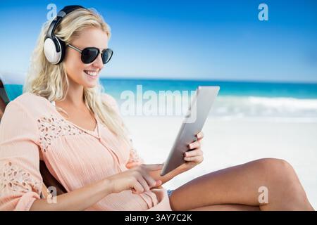 Frau, die auf Liegestuhl am Sandstrand sitzt, ein Tablet klopft und Kopfhörer und Sonnenbrille trägt Stockfoto
