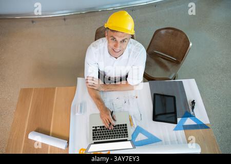 Mann mit gelbem Schutzhelm, der im Büro am Tisch sitzt, mit Laptop, Blaupausen und Tablet Stockfoto