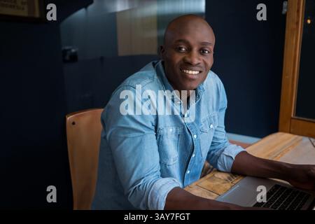 Junger Erwachsener Afroamerikaner mit Laptop in einem modernen Arbeitsbereich, mit Holztisch und Stuhl Stockfoto