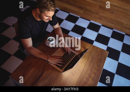 Mann tippt auf silbernem Laptop an Holztisch im Café, hält Keramikkkaffee, Kopierraum Stockfoto