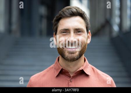 Ein fröhlicher Mann mit einem warmen Lächeln posiert vor einem Gebäude mit einer Steintreppe und strahlt Positivität aus. Stockfoto