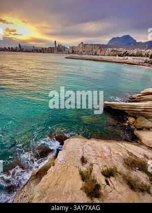 Ein goldener Sonnenuntergang umrahmt die Skyline einer mediterranen Stadt, während die Wellen sanft gegen zerklüftete Küstenfelsen krachen – ein Moment der Ruhe, in dem Natur ist Stockfoto