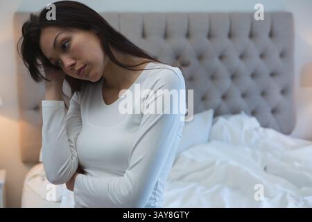 Junge asiatische Frau, die sich im Schlafzimmer mit dem Kopf auf die Hand lehnt, nachdenklich mit grauem Kopfteil, Kopierraum Stockfoto