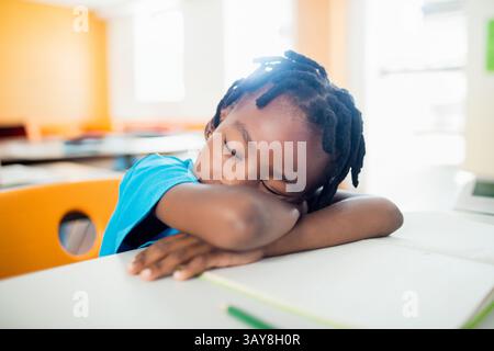Afroamerikanischer Junge, der im Klassenzimmer auf dem Schulpult schläft, mit Notizbuch und Bleistift Stockfoto