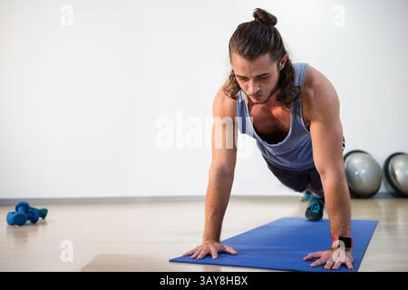 Mann, der Plank auf blauer Matte im Fitness Studio mit roter Smartwatch, blauen Kurzhanteln, Kopierraum spielt Stockfoto