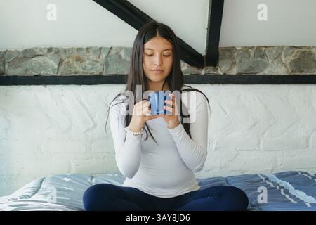Eine junge Frau sitzt bequem auf dem Bett mit einer Tasse in den Händen und nimmt sich einen Moment in einem ruhigen, gemütlichen Raum Stockfoto