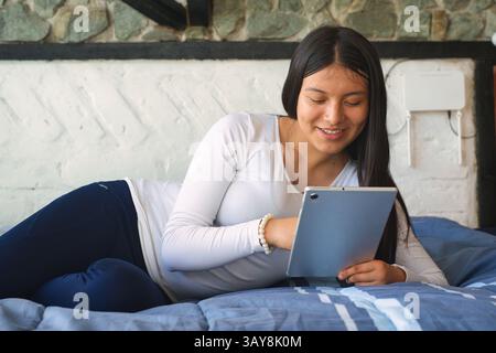 Eine junge Frau wird in einer stilvollen und einladenden Umgebung mit einem Tablet bequem auf ihrem Bett entspannt gesehen Stockfoto