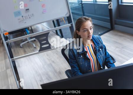 Frau, die Daten auf einem Computermonitor analysiert, während sie am modernen Schreibtisch mit Whiteboard-Ständer sitzt Stockfoto
