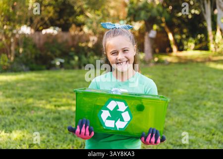 Mädchen mit grünem Recyclingbehälter im sonnendurchfluteten Garten, mit pinkfarbenen Gartenhandschuhen Stockfoto