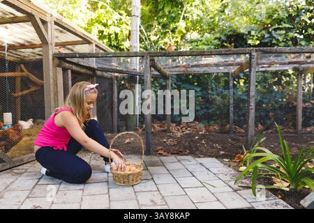 Kind kniet auf einer gepflasterten Terrasse im Hinterhof des Bauernhofes und sammelt Eier aus dem Hühnerstall in einen Korb Stockfoto