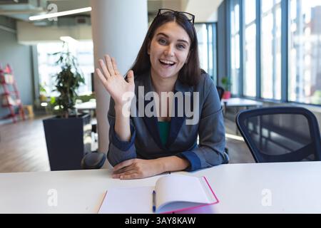 Frau, die am weißen Schreibtisch im Großraumbüro sitzt und mit rosa Notizbuch und blauem Stift winkt Stockfoto