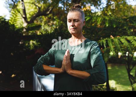Seniorin übt meditative Yoga-Pose auf Holzterrasse im Garten im Hinterhof, mit weißer Couch Stockfoto