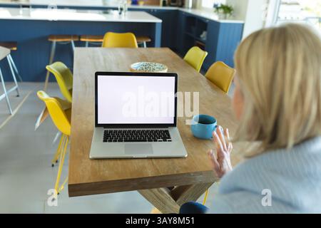 Ältere Frau, die Laptop und Gesten während eines Videogesprächs in der modernen Küche benutzt, mit blauem Becher, Schüssel Stockfoto