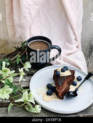 Ein Stück reichhaltiger Schokoladenkuchen mit Heidelbeeren und Sahne, serviert auf einer Keramikplatte neben einer schwarzen Tasse Kaffee. Stockfoto