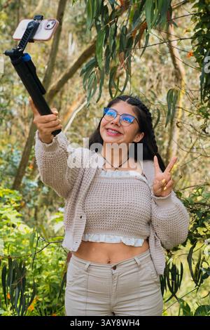 Eine freudige Frau nimmt mit ihrem Smartphone und einem Selfie-Stick ein lebhaftes Selfie in einer üppigen natürlichen Umgebung auf Stockfoto