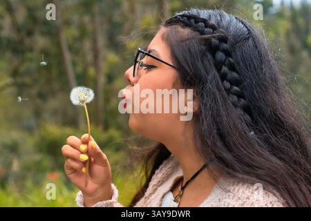 Eine junge Frau bläst sanft flauschige Löwenzahnsamen in die weiche, warme Brise in einer wunderschönen, ruhigen natürlichen Umgebung Stockfoto