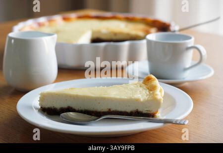 Stück selbstgemachter Käsekuchen auf einem weißen Teller auf Holztisch, eine Tasse schwarzen Kaffee mit Untertasse, eine Tasse Milch und eine Auflaufform im Hintergrund. Stockfoto