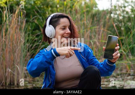 Eine junge Frau, die Kopfhörer trägt, genießt eine Musik-Streaming-App auf ihrem Telefon, zeigt auf den Bildschirm und lächelt Stockfoto