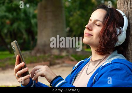 Entspannte junge Frau, die Musik mit schnurlosen Kopfhörern genießt und ihre Smartphone-App in einer friedlichen Parkumgebung durchsucht, während sie eine Pause einlegt, umgeben von n Stockfoto