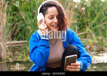 Glückliche junge Frau, die schnurlose Kopfhörer trägt, verwendet eine Musik-App auf ihrem Smartphone und genießt dabei die friedliche Atmosphäre einer natürlichen Umgebung Stockfoto