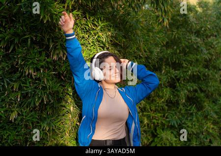 Die junge Frau, die Kopfhörer trägt, genießt Musik und tanzt mit geschlossenen Augen vor einer Bambushecke in einem sonnigen Park, hebt ihre Arme und smi Stockfoto