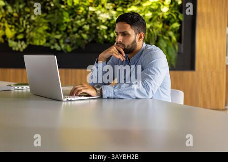 Junger Mann, der an silbernem Laptop arbeitet, am weißen Tisch im Büro, mit Pflanzenwand, Kopierraum Stockfoto