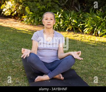 Ältere asiatische Chinesin meditiert auf schwarzer Yogamatte im grasbewachsenen Garten und übt Mudra-Pose Stockfoto