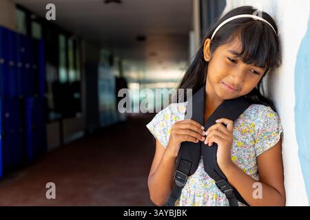 Kind Mädchen lehnt sich an die Wand im Schulflur, hält Rucksackgurte, Kopierraum Stockfoto