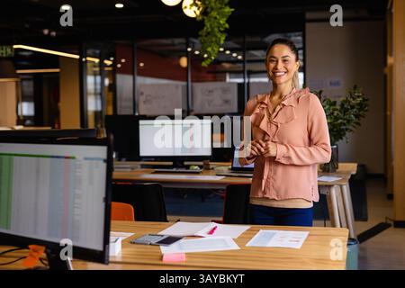 Frau, die im modernen Büro steht und lächelt, mit Computermonitor und Smartphone, Kopierraum Stockfoto