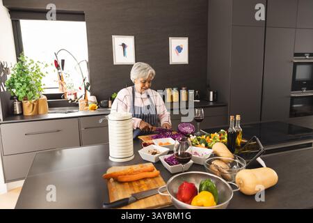 Ältere Frau, die Rotkohl in eine weiße Schüssel auf der Kücheninsel hackt, mit Holzschneidebrett Stockfoto