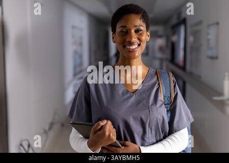 Afroamerikanerin, die im Krankenhausflur steht, Tablette hält und einen Rucksack trägt, lächelt Stockfoto