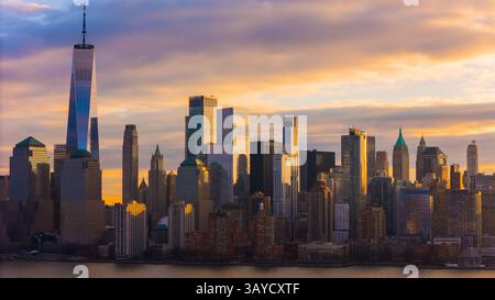 Die Skyline von Manhattan leuchtet unter einem dramatischen Sonnenuntergang, und das One World Trade Center erhebt sich über den eleganten Wolkenkratzern und den urbanen Silhouetten der Stadt. Stockfoto