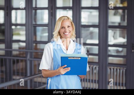 Junge Erwachsene Frau, die im Flur des Büros steht und hellblaues Klemmbrett mit beiden Händen hält, lächelnd Stockfoto