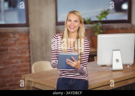 Frau, die auf hölzernem Schreibtischrand in einem modernen Bürobereich sitzt und ein Tablet in der Nähe von Monitor und Pflanze hält Stockfoto