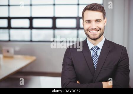 Geschäftsmann, der am Schreibtisch steht und die Arme im Büro überquert, mit schwarzem Anzug gestreiftem Krawattenkrawatte, Laptop und Fenster Stockfoto