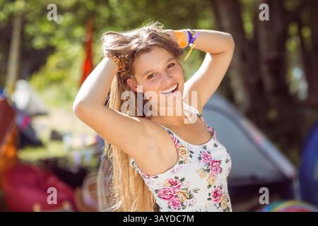 Junge Frau, die Haare auf dem Waldcamping mit verschwommenen Zelten repariert, mit Blumenoberteil und Armbändern Stockfoto
