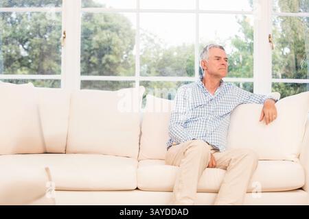 Seniorenmann sitzt auf weißem Sofa im Wohnzimmer, blickt aus dem Fenster mit Zimmerpflanze, Kopierraum Stockfoto