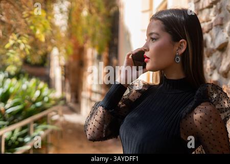 Eine junge Geschäftsfrau mit rotem Lippenstift und elegantem schwarzem Kleid telefoniert in einer eleganten, urbanen Umgebung, umgeben von Grün und ston Stockfoto