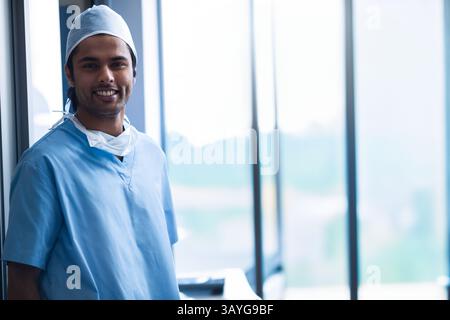 Junger asiatischer männlicher Arzt, der in der Krankenhaushalle an den Fenstern steht, Schrubben, Kappe, Maske, Kopierraum trägt Stockfoto