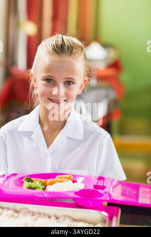 Das Schulmädchen lächelt und hält ein rosafarbenes Tablett mit Reis Karotten Brokkoli in der Cafeteria und genießt ein Mittagessen Stockfoto
