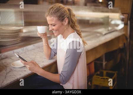 Frau, die auf der Marmorcafé-Theke sitzt und ihr Smartphone überprüft, weiße Kaffeetasse mit Metallregal hält Stockfoto