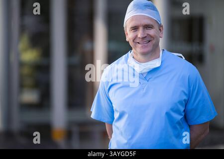 Leitender Chirurg mit blauen Peelings und Maske vor dem Krankenhauseingang stehend, lächelnd, Kopierraum Stockfoto