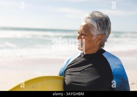 Seniorin steht mit gelbem Surfbrett am Strand in schwarzem oder blauem Neoprenanzug und genießt die Meeresbrise Stockfoto