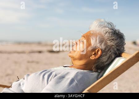 Ältere Frau, die sich auf einem Liegestuhl mit gestreiftem Kissen am Strand liegend, sonnig, Kopierraum Stockfoto