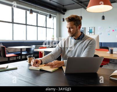 Mann im mittleren Erwachsenenalter, der in Spiralnotizbuch auf dem Schreibtisch im modernen Großraumbüro schreibt, mit Laptop Stockfoto